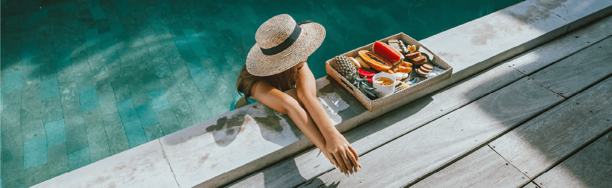 Woman in straw hat relaxing by turquoise pool with tropical breakfast tray featuring fresh fruits and tea